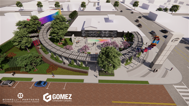 Wide aerial view of an entire memorial site including a curved entry structure, landscaped walkways, colorful glass elements, trees, and the central plaza with a rainbow-colored rectangular installation. Surrounding streets, sidewalks, and buildings frame the space.