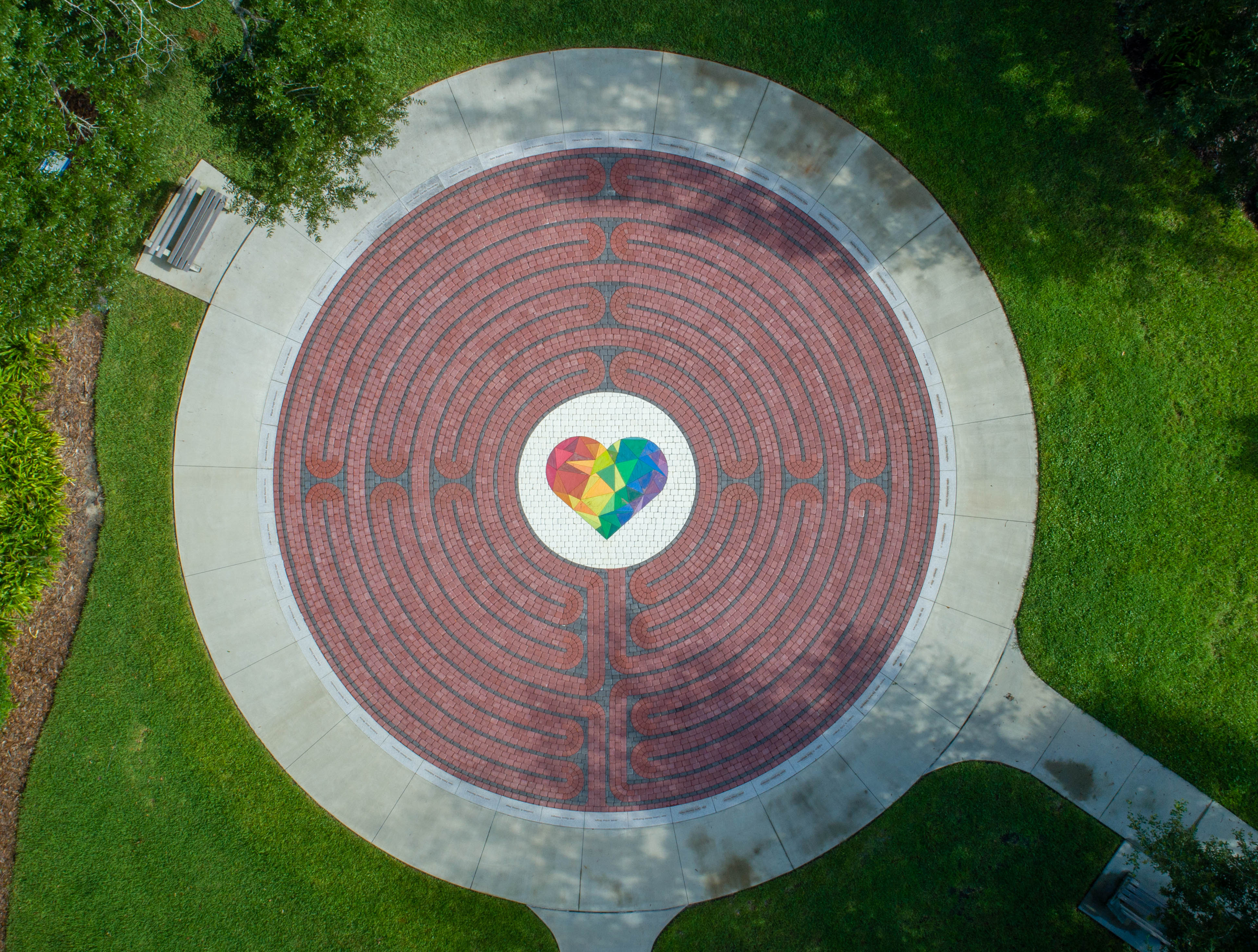Aerial view of labyrinth. Green space outside of the circle, first outer layer is a basic grey path. Next layer in is the maze part with the pavers. The middle is a prismatic, rainbow heart. 